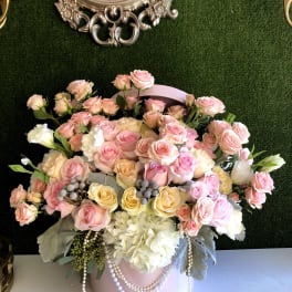 Large pastel arrangement of pink and yellow roses with white hydrangeas in a pink hatbox with pearls