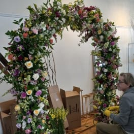 Large floral arch covered in mixed blooms with a person arranging it nearby