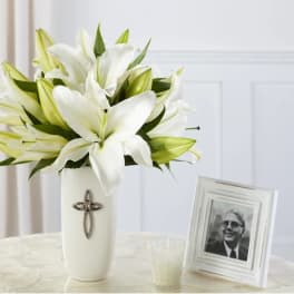 White lily bouquet in a white vase with silver cross beside framed photo and candle