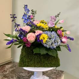 Mixed bouquet with roses, hydrangea, daisies, and purple blooms in a moss-covered container