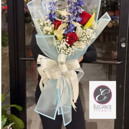 Hand-tied bouquet of red roses, white calla lilies, and purple flowers wrapped in pale blue paper