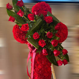 Standing red floral cross with red roses, large green leaves, and ribbon on a wooden easel.