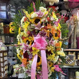 Large standing floral spray with lilies, sunflowers, roses, and a pink ribbon
