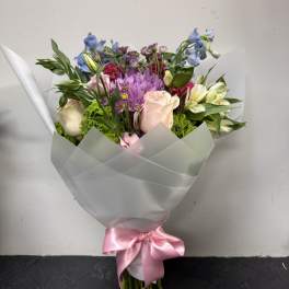 Mixed bouquet of pink, purple, blue, and white flowers wrapped in white paper with a pink ribbon