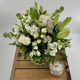 White and cream roses, lilies, and stock in a clear glass vase with a candle beside on a wooden crate