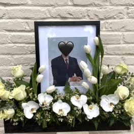 White floral tribute around a framed portrait in a black container.