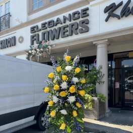 Standing floral spray with yellow roses, white blooms, and purple accents