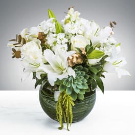 Low round arrangement of white lilies and roses with succulents in a green glass bowl vase.