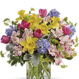 Mixed bouquet of yellow daffodils, pink tulips, and purple flowers in a glass vase
