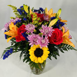 Bright mixed bouquet with sunflower, red roses, orange lilies, and purple mums in a clear glass vase