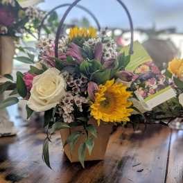 Mixed arrangement with sunflower, white rose, and pink blooms in a small kraft pot with handle.
