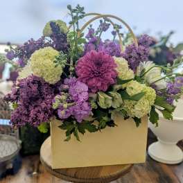Purple and green mixed flower arrangement in a cream tote-style box on a wooden table