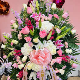 Tall standing spray of pink and white roses, lilies, and carnations with a large pink bow.