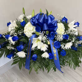 Large blue and white funeral spray with blue roses, white lilies, and a blue bow on a bed of dark foliage.