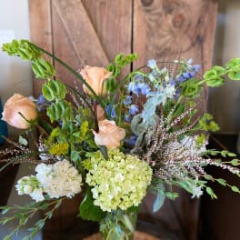 Tall vase arrangement with peach roses, green hydrangeas, bells of Ireland, and mixed accent flowers