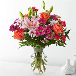 Mixed bouquet of pink lilies, orange roses, and magenta daisies in a glass vase