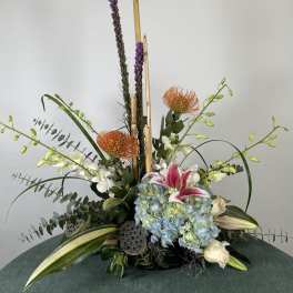 Tall floral arrangement with blue hydrangeas, lilies, and orange pincushion blooms