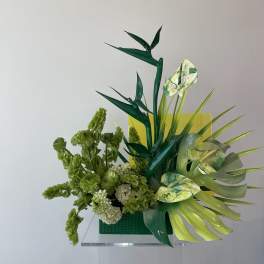 Green floral arrangement with tropical leaves and pale blooms in a square vase