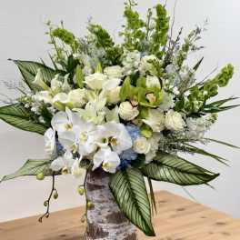 Tall arrangement of white orchids, roses, lilies, and green orchids in a rustic cylinder vase