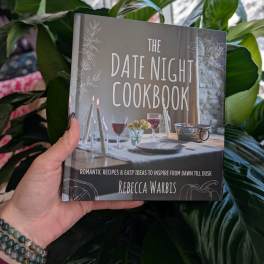 Hand holding a cookbook in front of large green houseplants