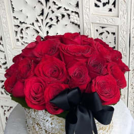 Red roses arranged in a round gold-patterned box with a black ribbon
