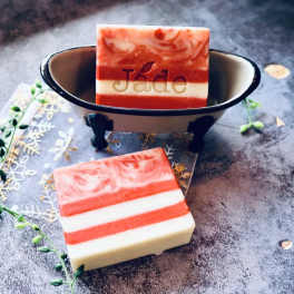 Striped red and white bar soaps, one in a small metal bathtub dish on a textured surface.