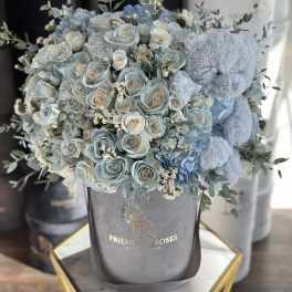 Blue roses and hydrangeas in a gray hatbox with a teddy bear