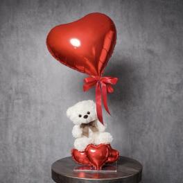 White teddy bear with bow sitting among red heart balloons and a large red heart balloon above.