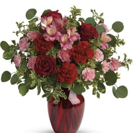 Red and pink roses, carnations, and alstroemeria arranged in a red glass vase