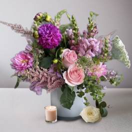 Pink and purple flower arrangement in a white vase with a candle and rose