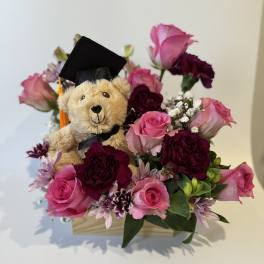 Teddy bear in a graduation cap surrounded by pink and purple flowers in a wooden box