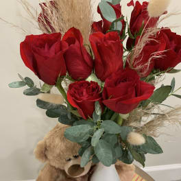 Red roses in a white vase with a teddy bear beside them