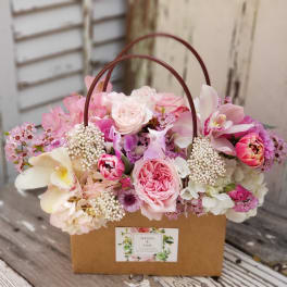 Pink and white roses, orchids, and mixed blooms arranged in a kraft paper tote with handles.