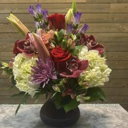 Mixed bouquet with roses, lilies, hydrangeas, and purple blooms in a dark vase
