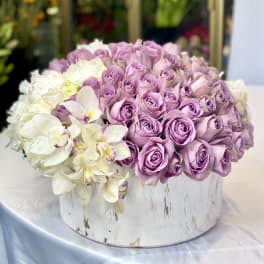 Lavender roses and white orchids arranged in a round white box