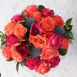 Round bouquet of bright orange and hot pink roses with dark purple calla lilies and blue thistle blooms.