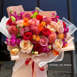 Large bouquet of red, pink, and peach flowers wrapped in blush paper