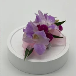 Lavender orchid corsage with bright purple accents and pink ribbon on a white round base.