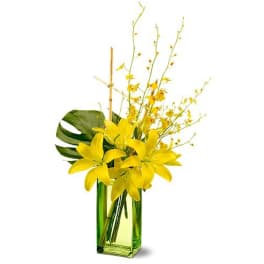 Tall arrangement of yellow lilies and small yellow orchids in a green glass rectangular vase with a large leaf