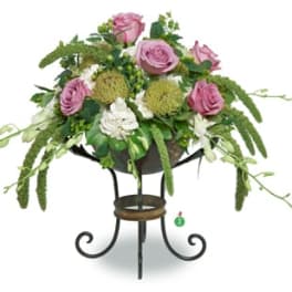 Pink roses, green mums, and white orchids arranged in a dark metal pedestal bowl on a scroll stand
