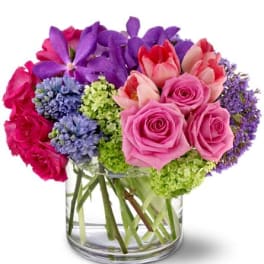 Low glass vase of pink roses, red-tipped tulips, purple orchids, blue hyacinths, and green hydrangea.