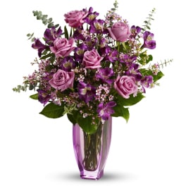 Tall arrangement of lavender roses and purple flowers in a pink glass vase