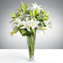 Tall arrangement of white lilies and baby’s breath in a clear vase with variegated green leaves and green ornaments