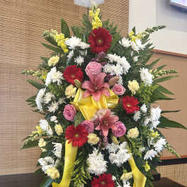 Standing floral wreath with pink lilies, red gerberas, white daisies, and yellow ribbon