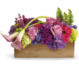 Low wooden box arrangement of pink roses, purple blooms, and pale calla lilies.