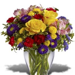 Bright mixed bouquet with yellow roses, red carnations, and purple blooms in a clear glass vase