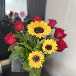 Arrangement of bright yellow sunflowers and red roses in a clear glass vase on a dark table.