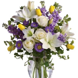 Mixed bouquet of white lilies and roses with purple flowers and yellow tulips in a clear glass vase