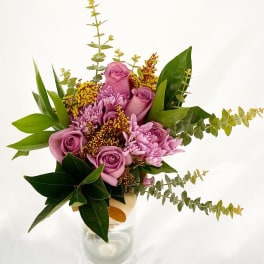 Tall bouquet of mauve roses and lavender mums with yellow filler in a clear glass vase