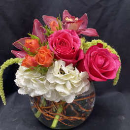 Short arrangement of hot pink roses, orange spray roses, orchids and white hydrangea in a glass bowl vase